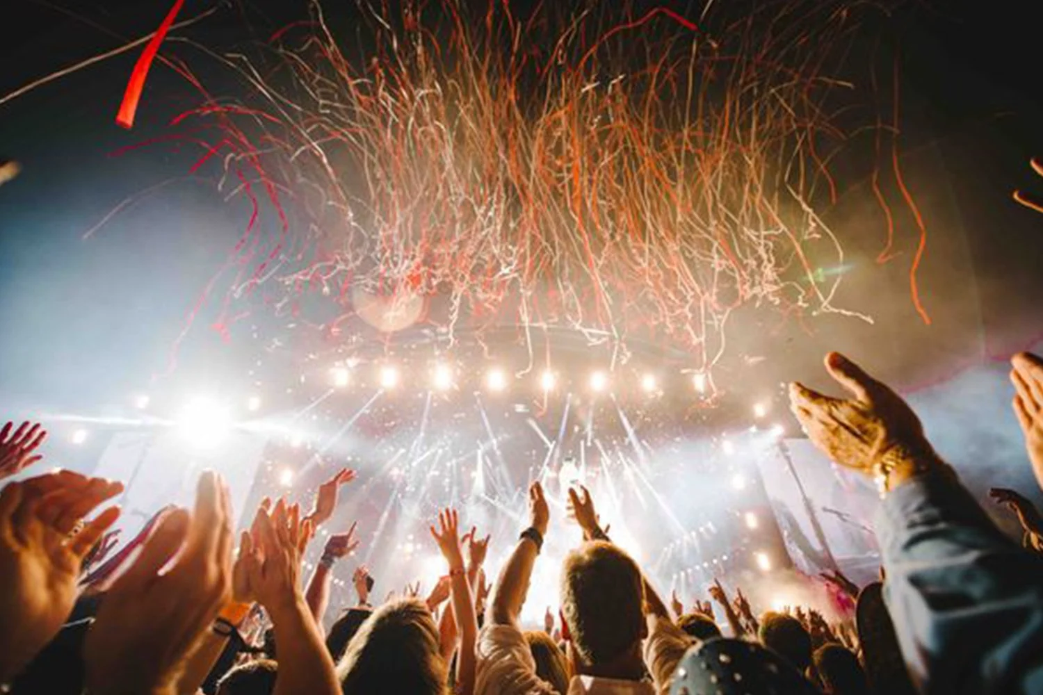 Crowd at concert with fireworks in the sky, people raising hands, and bright stage lights.