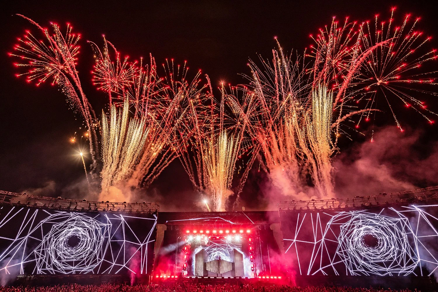 Fireworks explode above a stage at a concert or festival with geometric light designs.