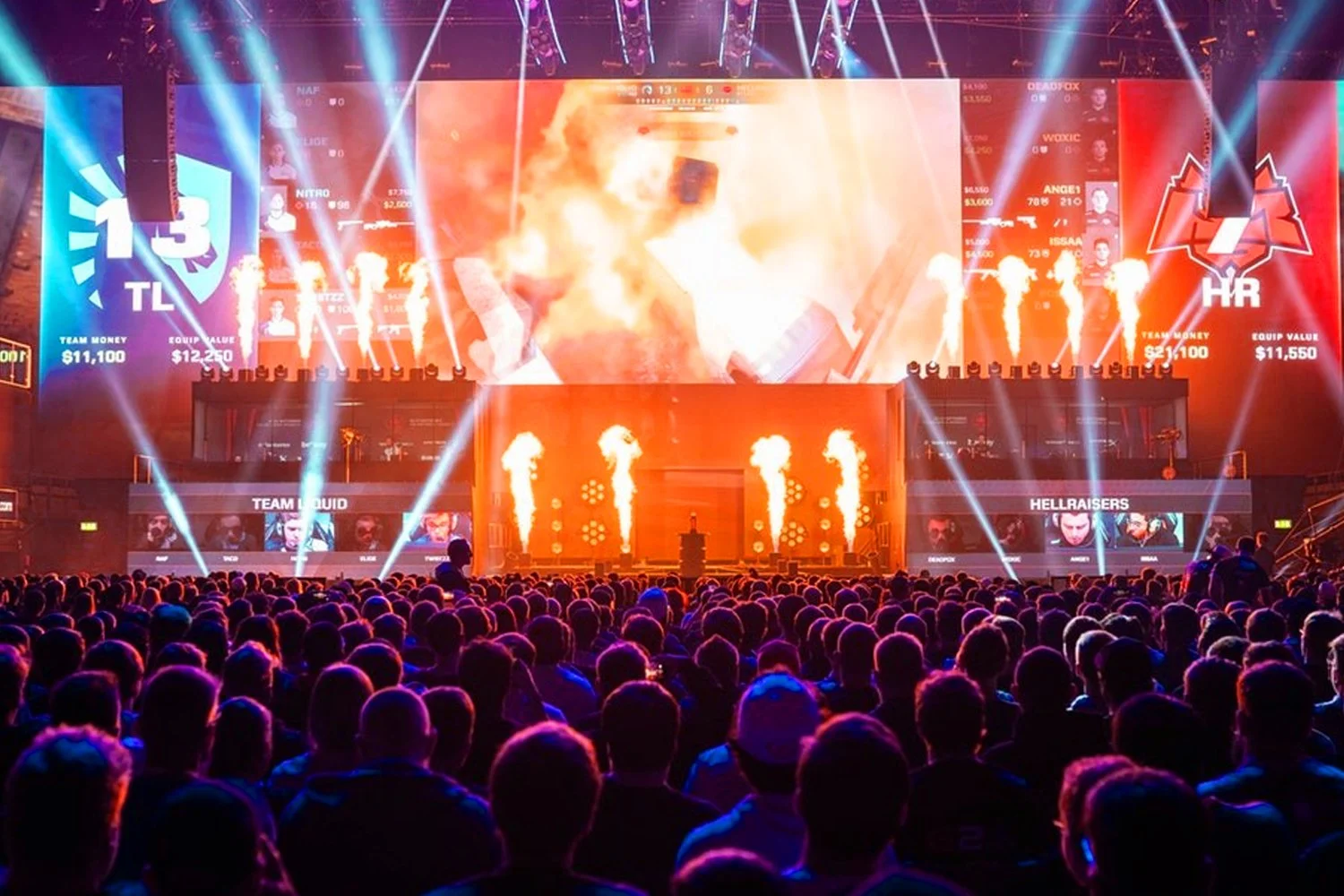 A large crowd watching an eSports event with bright lights, flames, and a giant screen showing team scores and players on stage.