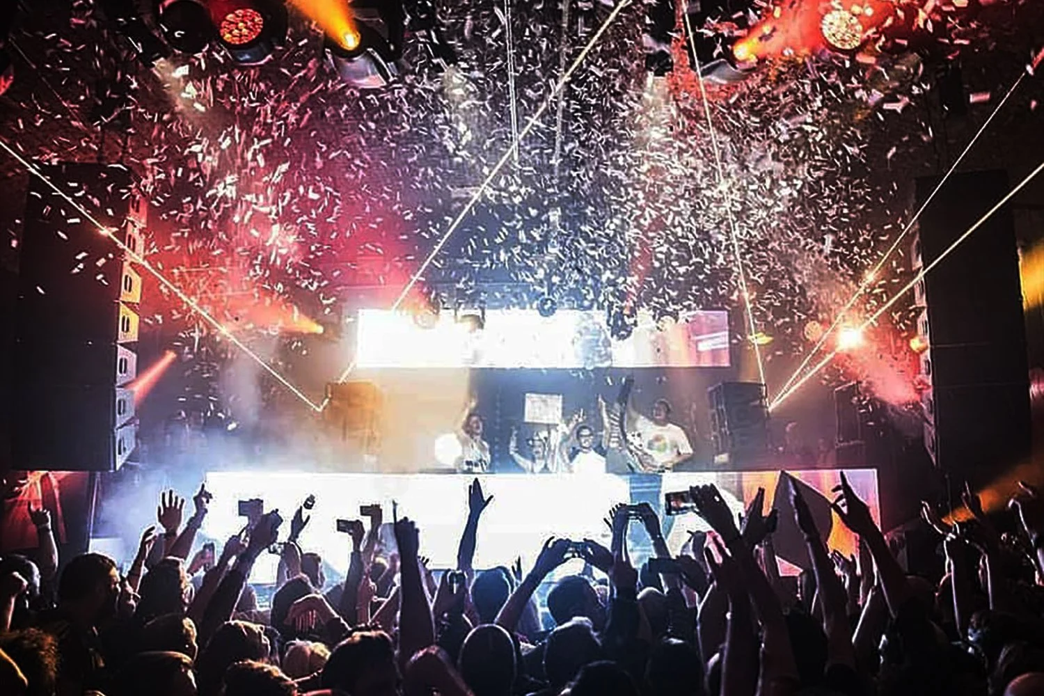 Crowd at a concert with hands raised, stage with performers, colorful stage lights, fog, and confetti falling from the ceiling.