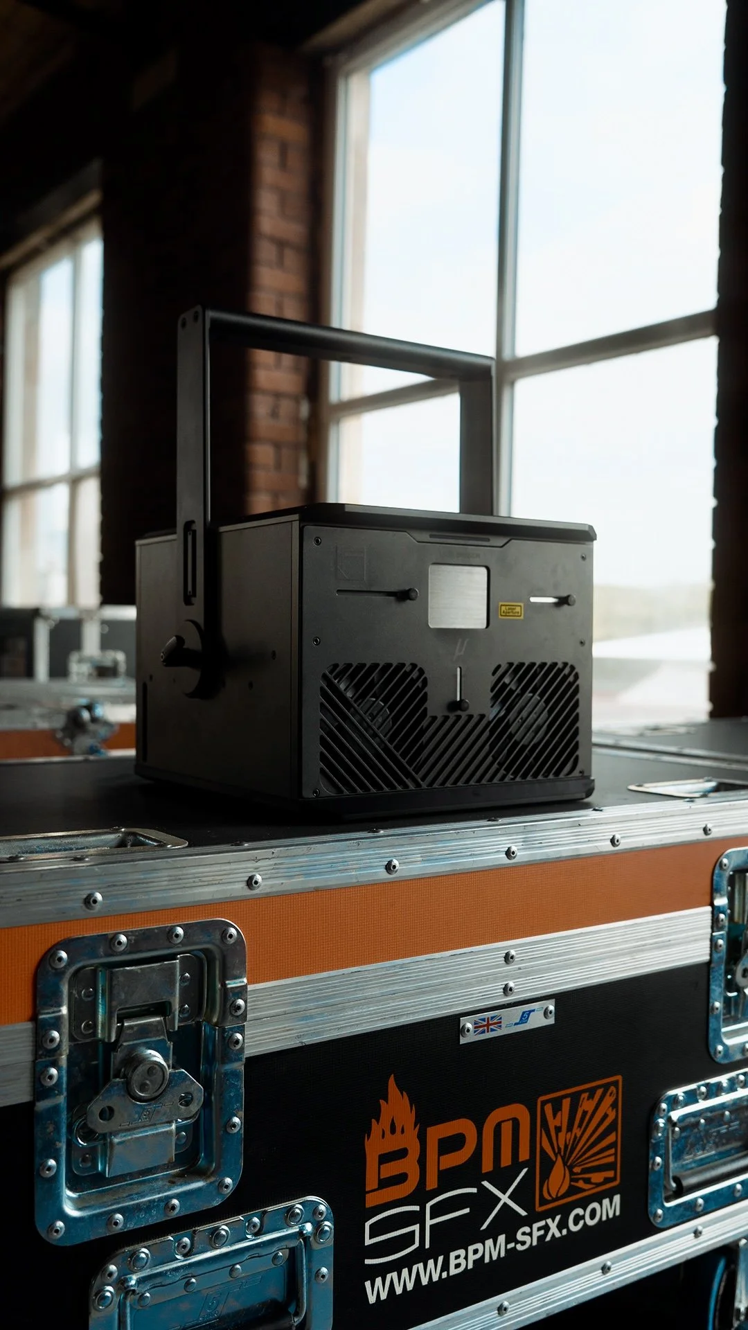 A black electronic device with a handle on top, placed on a flight case with orange and black colors, and the logo 'BPM SFX' and website 'www.bpm-sfx.com' on it. The background shows large windows letting in natural light.