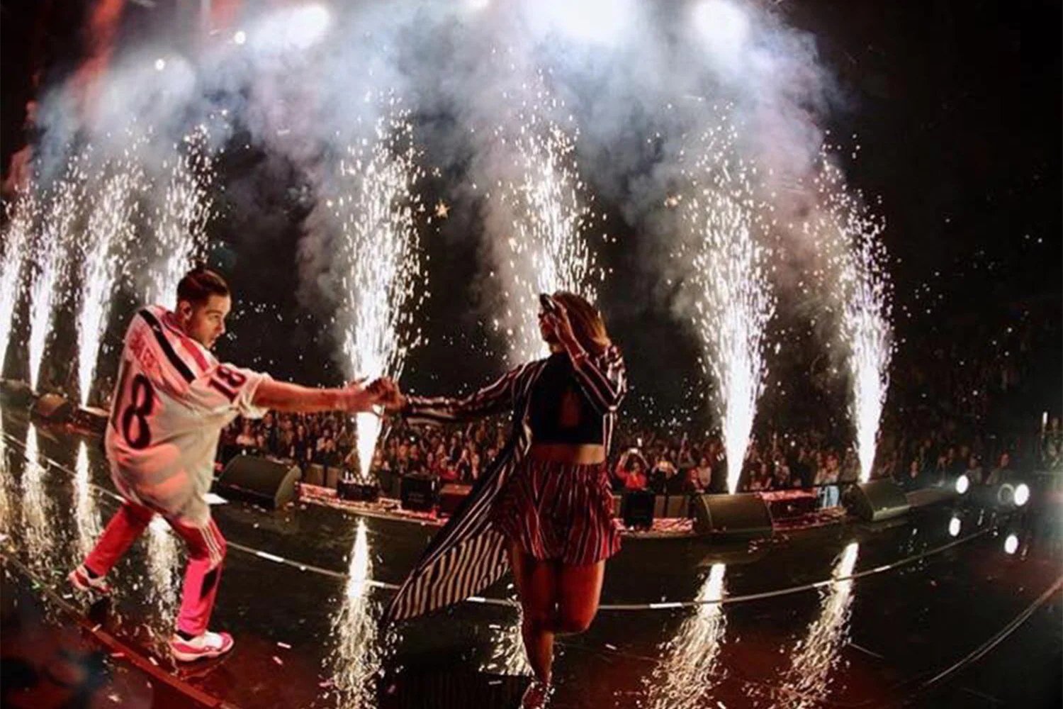 Two performers on stage holding hands, surrounded by fireworks and a crowd at a lively concert or event.