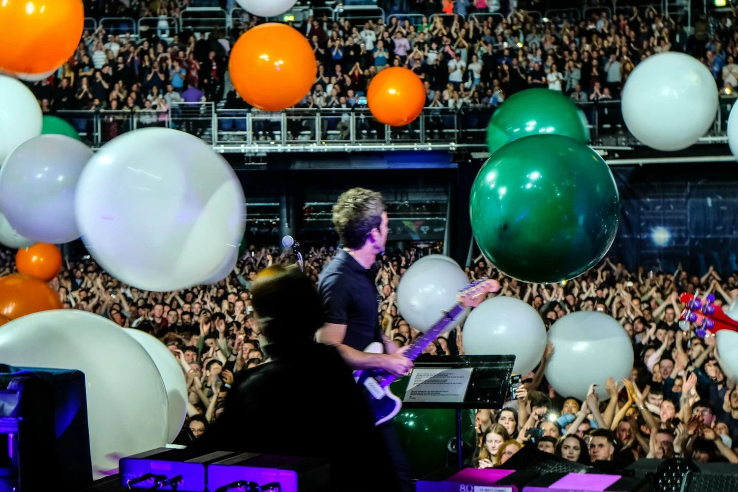 Concert scene with a musician playing guitar on stage, surrounded by large white, green, orange, and purple balloons, and a large crowd with hands raised in the background.