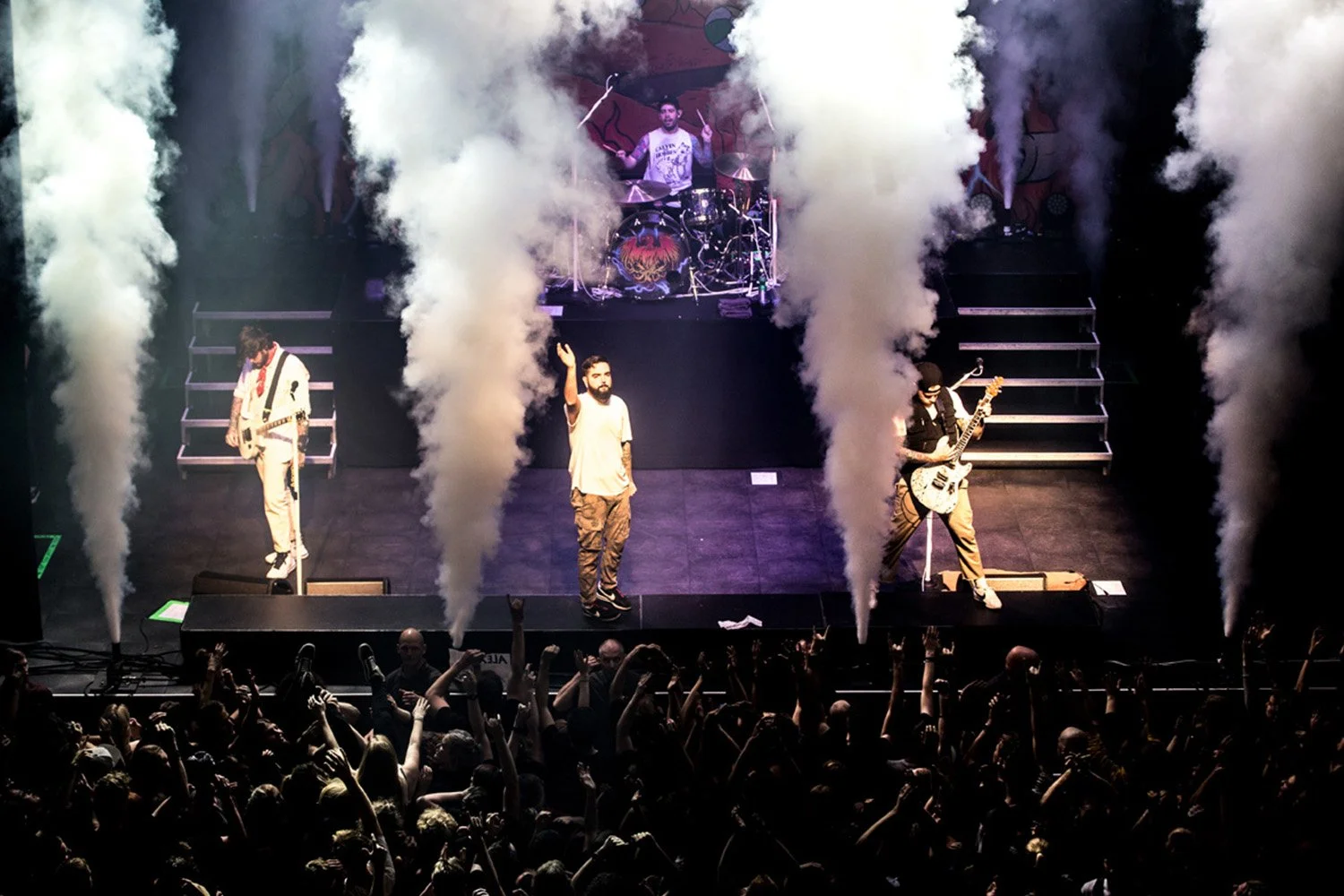 A live concert performance with three band members on stage, a drummer in the background, and colorful smoke effects. The crowd is cheering and raising their hands in front of the stage.