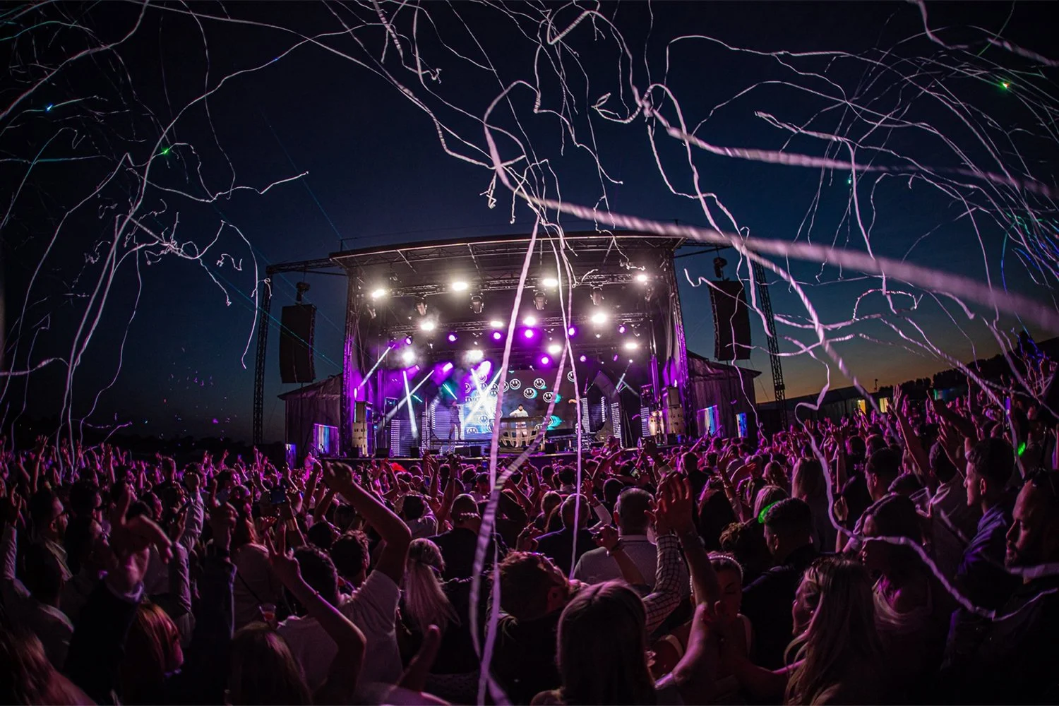 A large outdoor concert at night with a crowd of people dancing and cheering, a stage with bright purple and white lights, and streamers flying through the air.