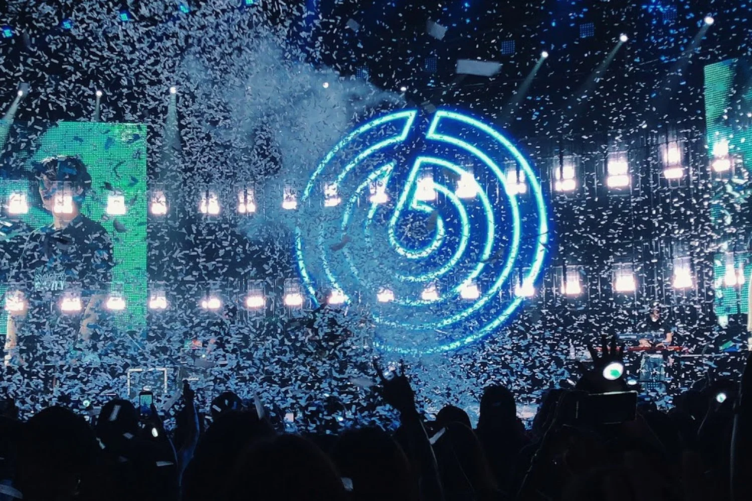 A concert scene with a crowd, falling confetti, bright blue neon spiral light, and large digital screens with a performer.