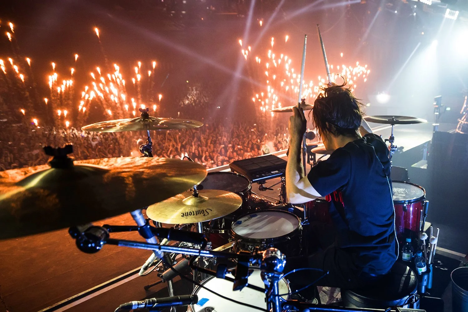 A drummer on stage playing drums during a concert, with fireworks illuminating the crowd in the background.