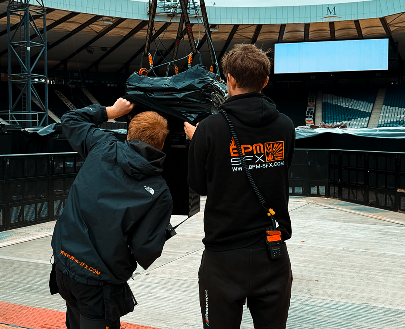 Two crew members working together to set up equipment at a stadium, with one person facing away and the other crouching, both wearing black jackets with logos, and the stadium appears to be an outdoor arena with empty seats and a large digital screen.