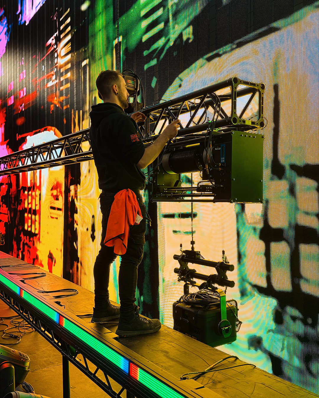 Technician working on a large LED screen with colorful digital graphics in the background, standing on a platform with tools and equipment around.