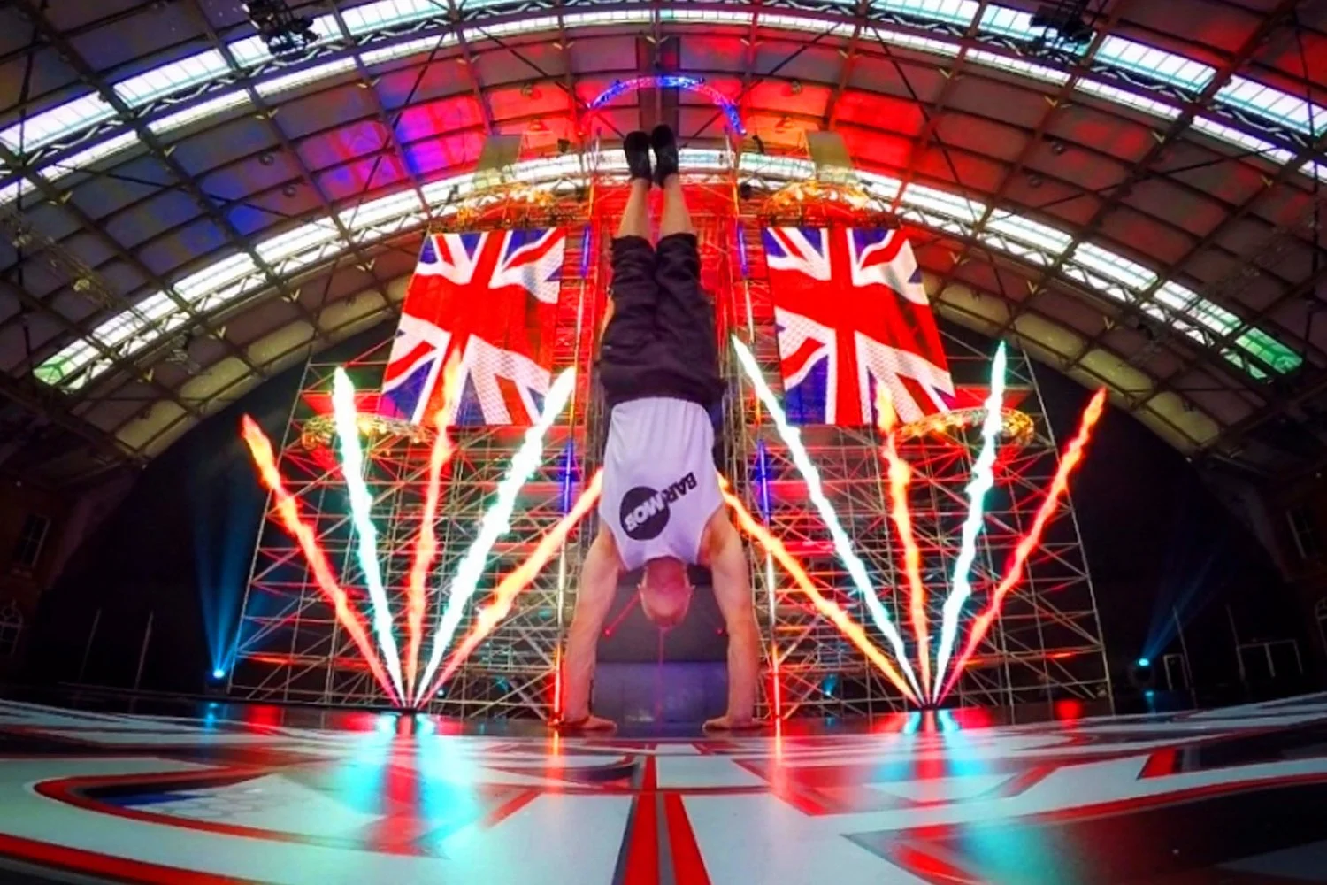 A performer doing a handstand at an indoor event with a stage backdrop of large British flags and fireworks lighting effects.