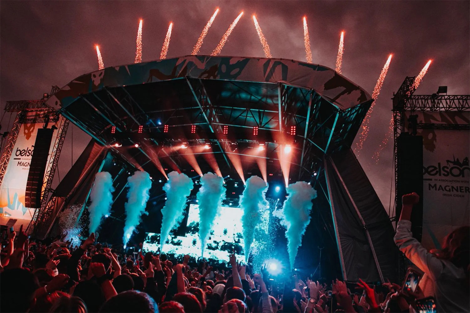 Concert stage with colorful lighting, smoke effects, and fireworks in the sky, surrounded by an audience.