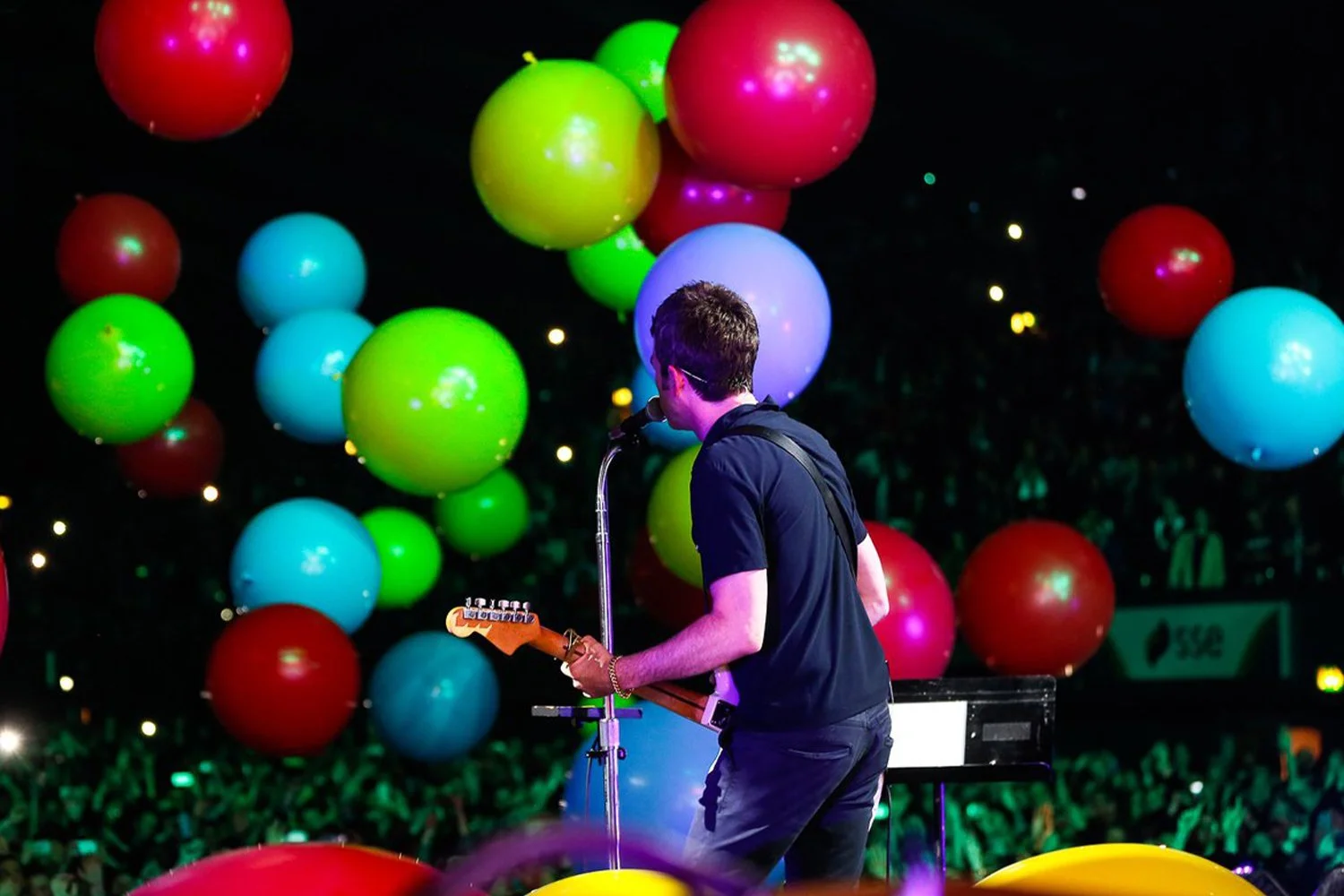 A musician on stage playing an electric guitar with colorful balloons floating behind him, and a crowd of people in the background.