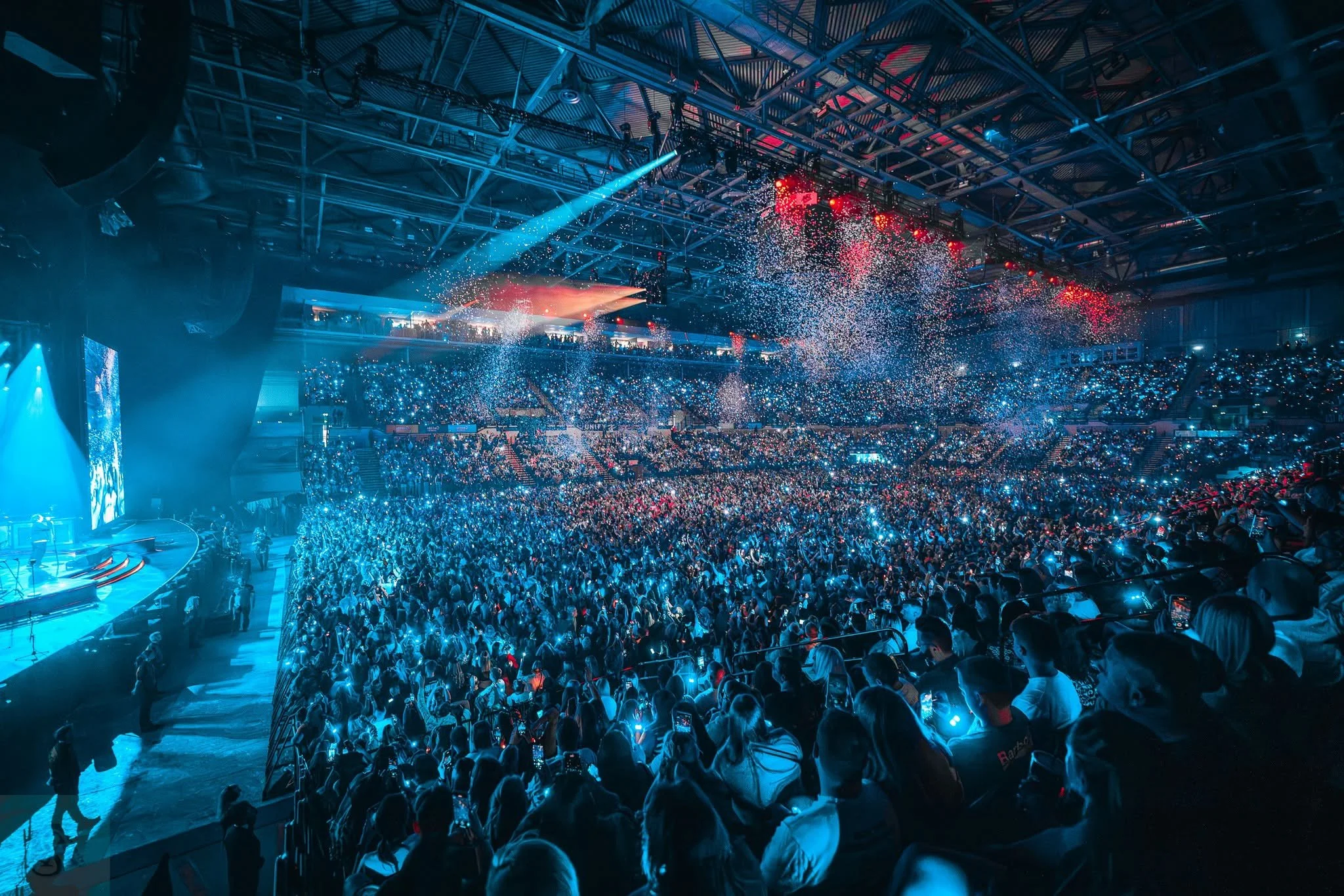 Audience in a large indoor arena with blue and red stage lighting, confetti, and many people standing, some taking photos or videos of an event.