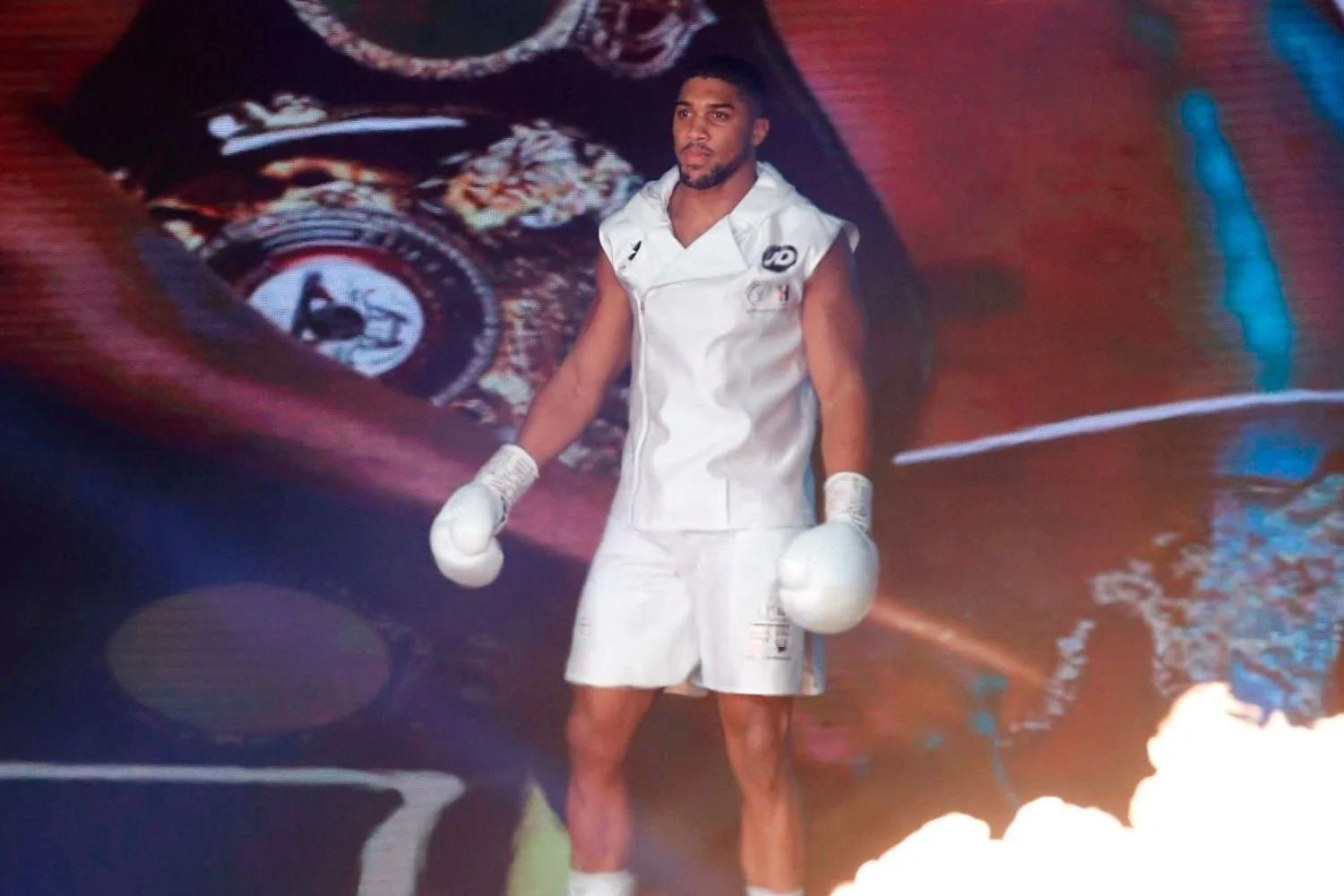 A male boxer dressed in white boxing gear standing in a boxing ring, with a large colorful graphic design projected behind him.