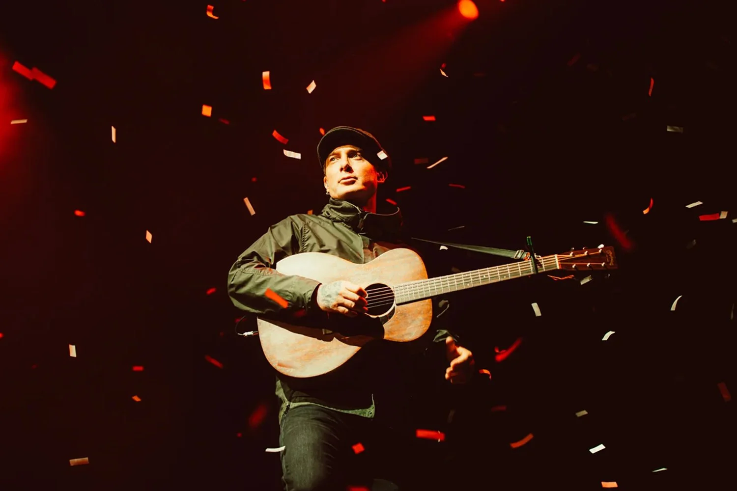 A man in dark clothing and a cap playing an acoustic guitar on stage with red and white confetti falling around him, illuminated by stage lighting.