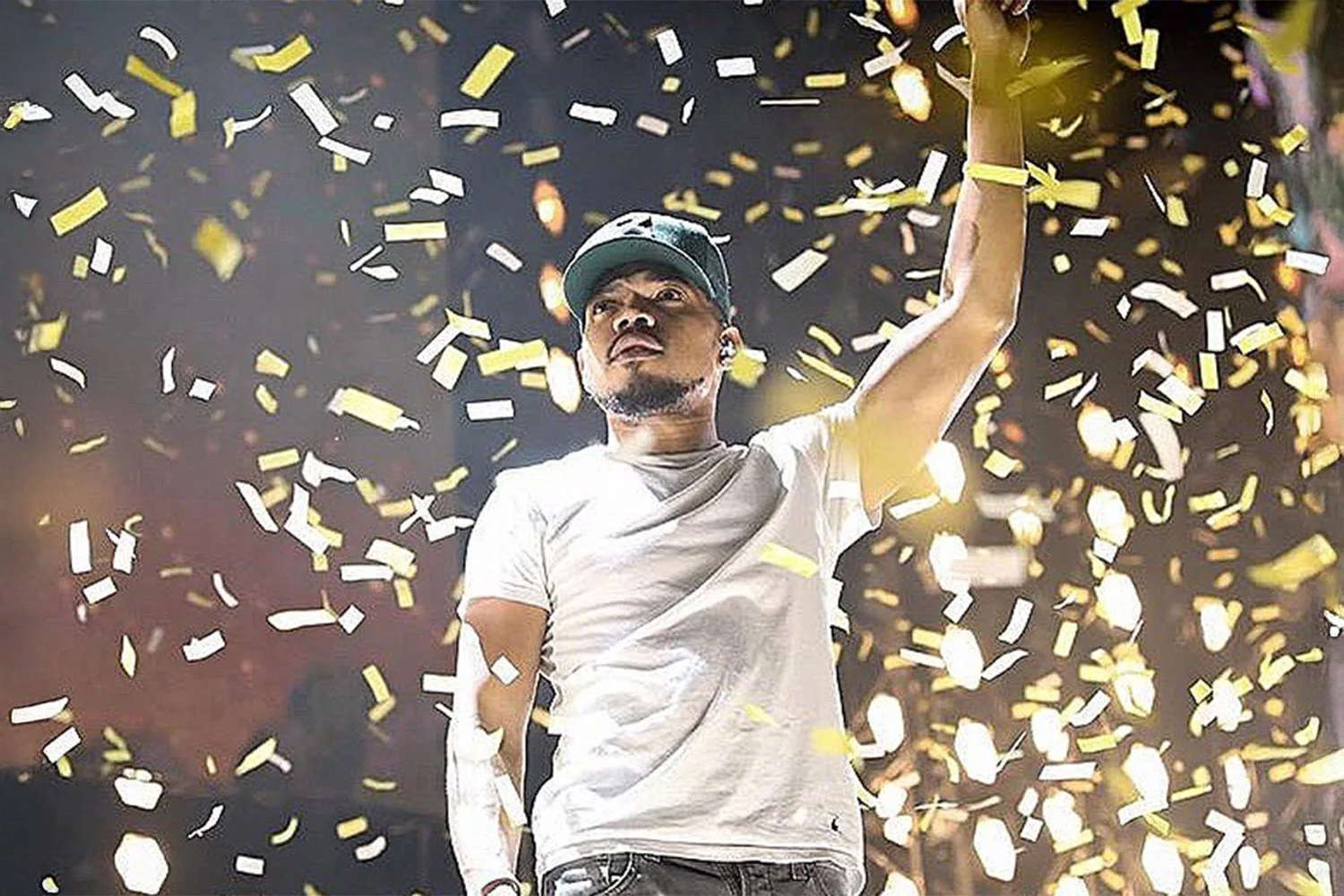 A man wearing a baseball cap and white t-shirt stands underneath falling gold and white confetti, appearing triumphant during a celebration or concert event.