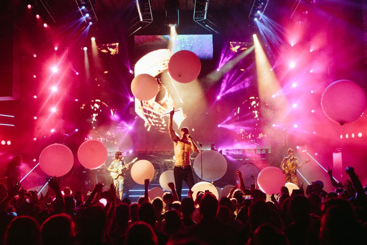 Live concert with performers on stage, colorful lighting, large pink balloons, audience with raised hands, and a big screen behind the stage showing a close-up of a performer.