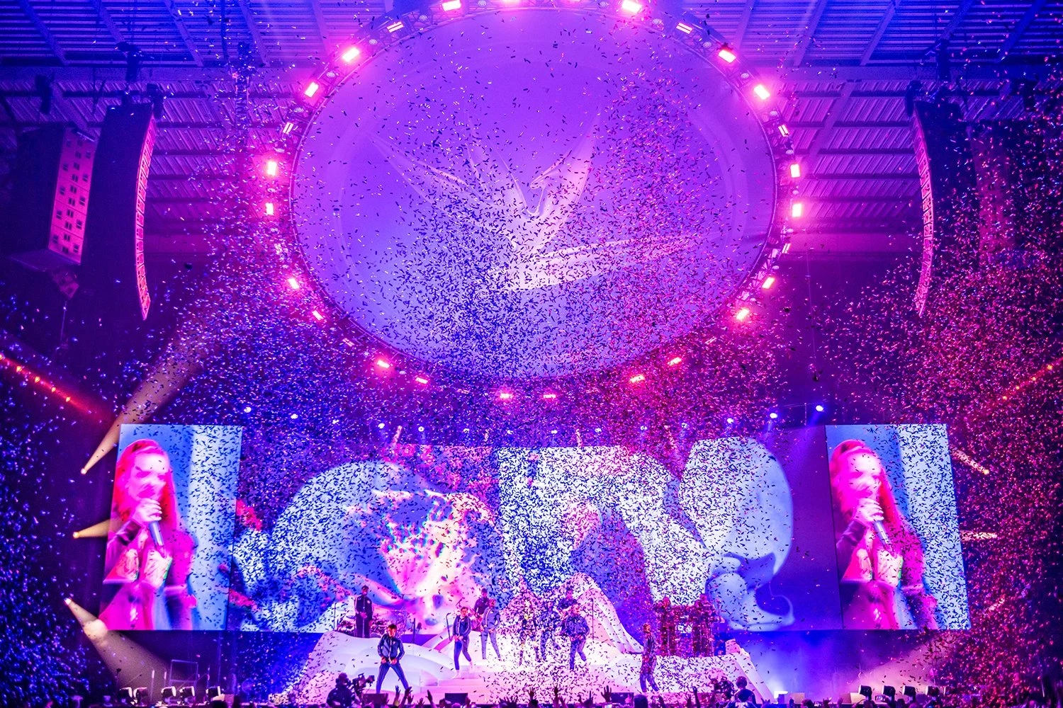 A lively concert scene with performers on stage, colorful lighting, large screens displaying images, and confetti falling from above.