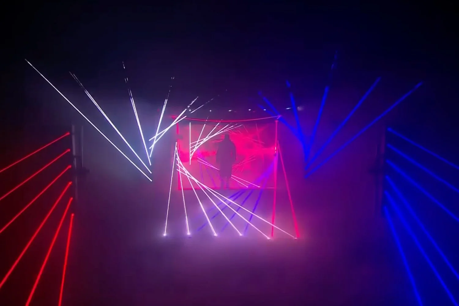 Person standing in the center of a dark room surrounded by colorful laser light effects in red, white, and blue, creating a tunnel-like visual display.