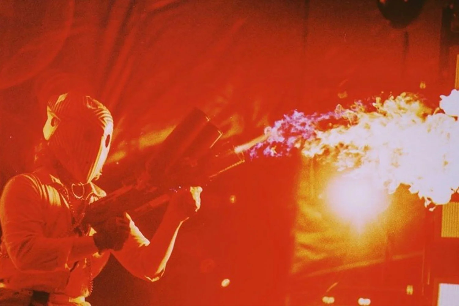 A person wearing a hood and mask, holding a flame-thrower or similar device, with a large burst of fire and flames shooting out, illuminated by red and orange lighting.