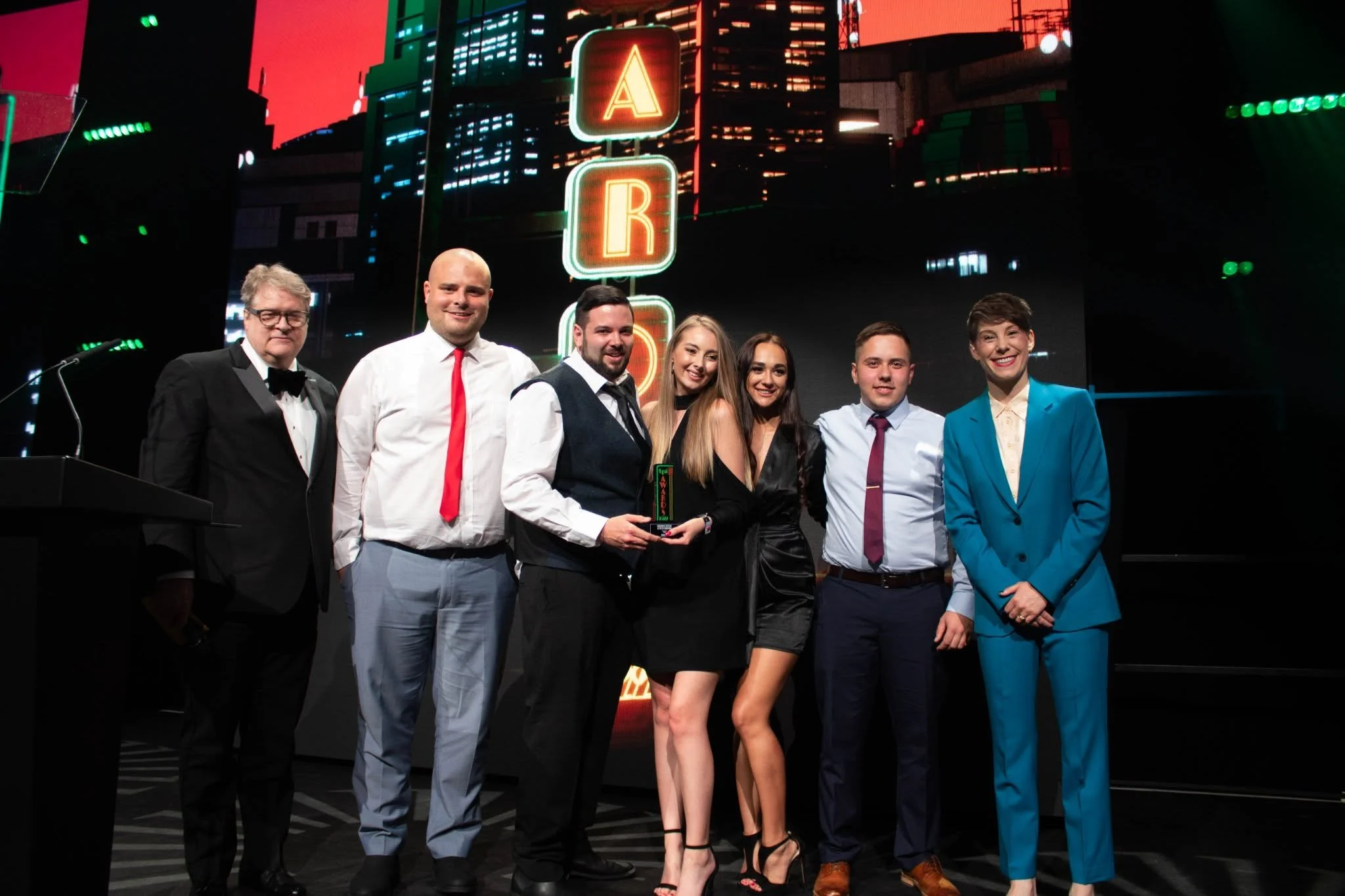 A group of seven people dressed in formal attire standing on stage in front of a backdrop with neon signs spelling 'A R E' against a cityscape background. The group appears to be at an award ceremony or event.