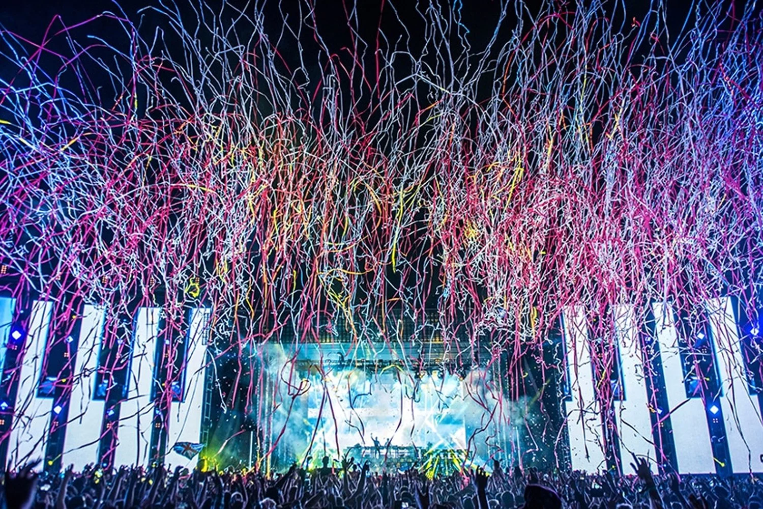 A concert stage with a crowd, colorful streamers and lights in the air, illuminated screens, and neon lighting effects.