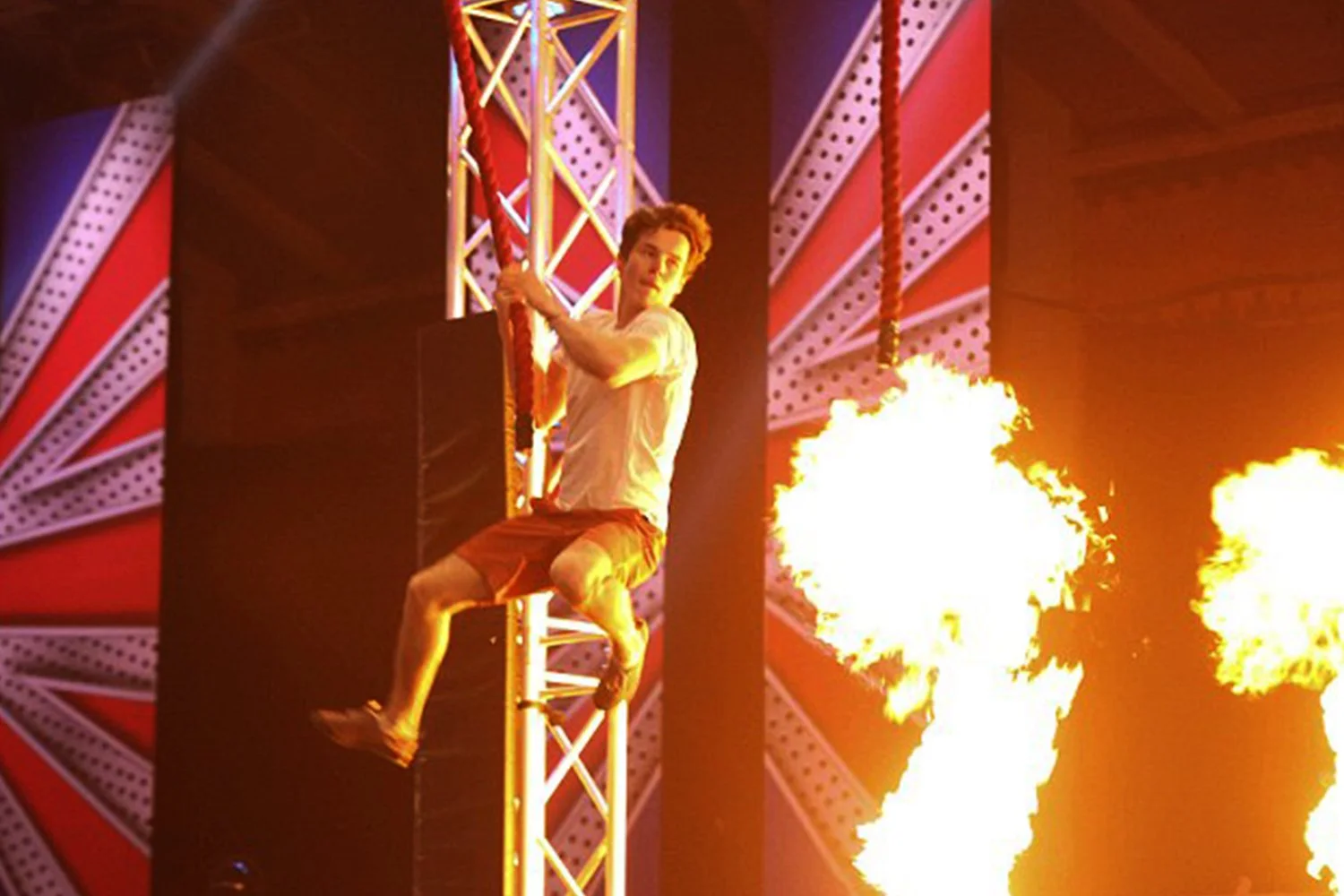 A man is hanging from a fire rope inside a circus or performance venue with fire flames in the background.