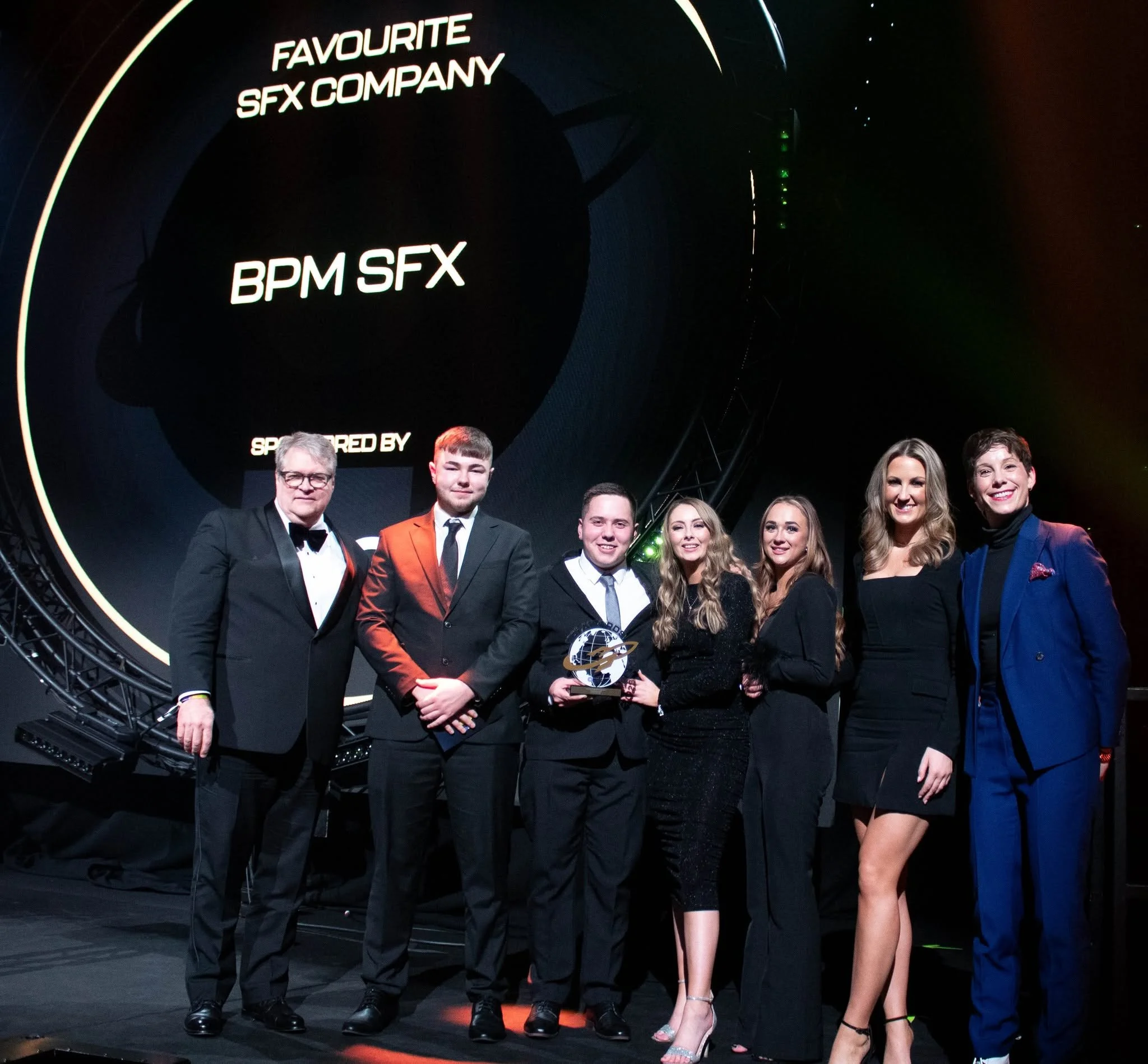 Group of people dressed in formal attire standing on stage at an awards event, with a large screen behind them displaying text about a company.