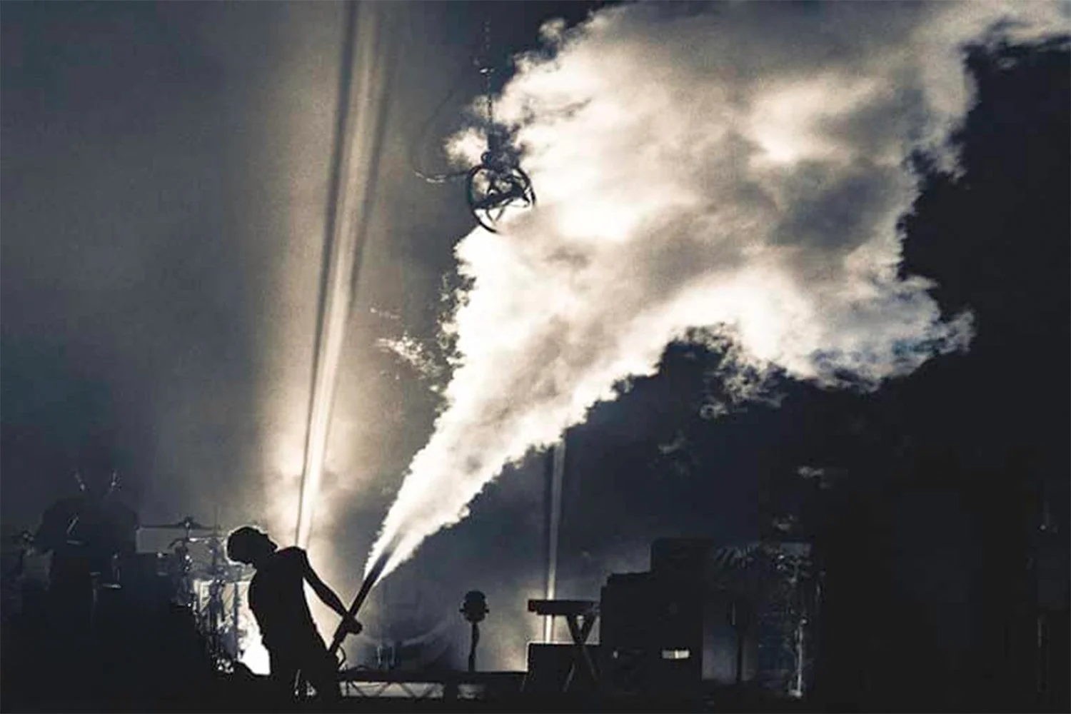 A person on stage holding a smoking pyrotechnic device during a concert or performance, with smoke and laser lights in the background.