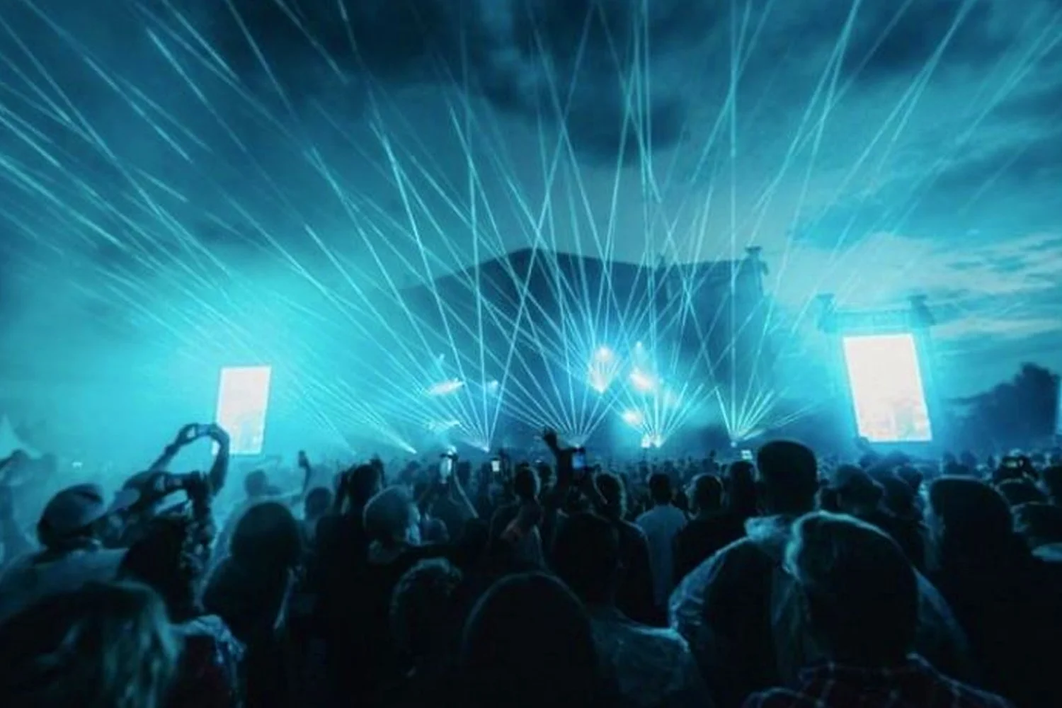 Crowd attending a concert or festival with blue laser lights and large screens.