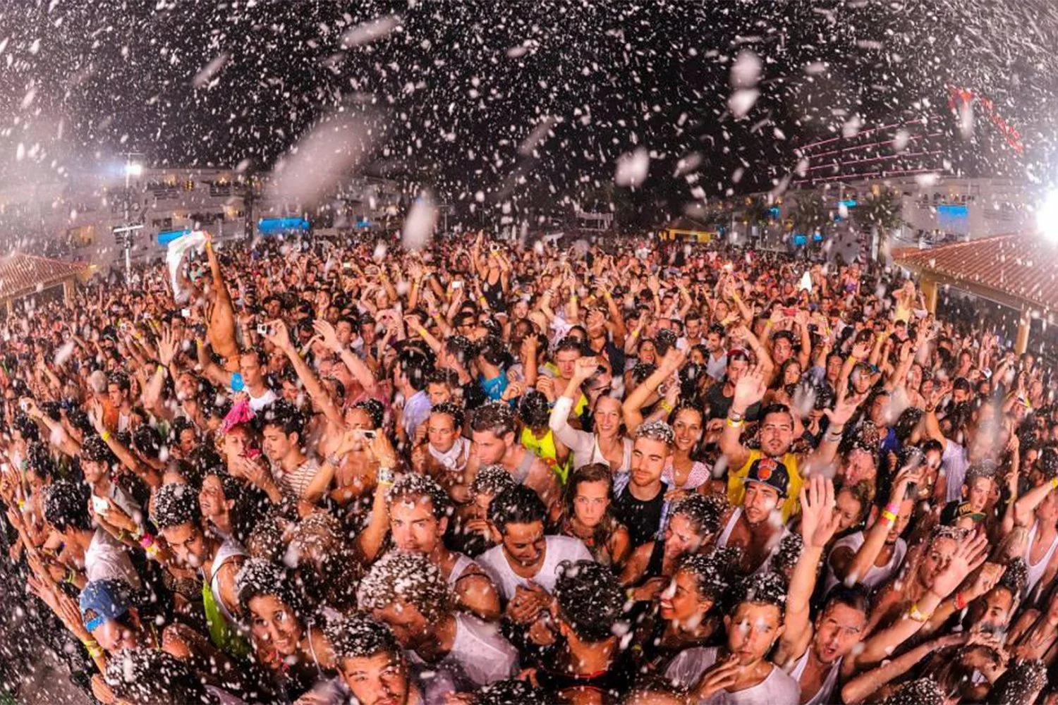 Crowd of people at a concert or festival, with confetti falling and many raising their hands, nighttime setting with bright lights.