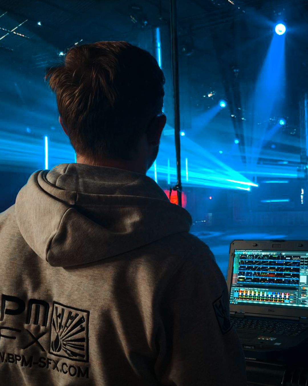 A person in a hoodie working with a laptop, producing electronic music in a dark room with blue stage lighting and laser effects.