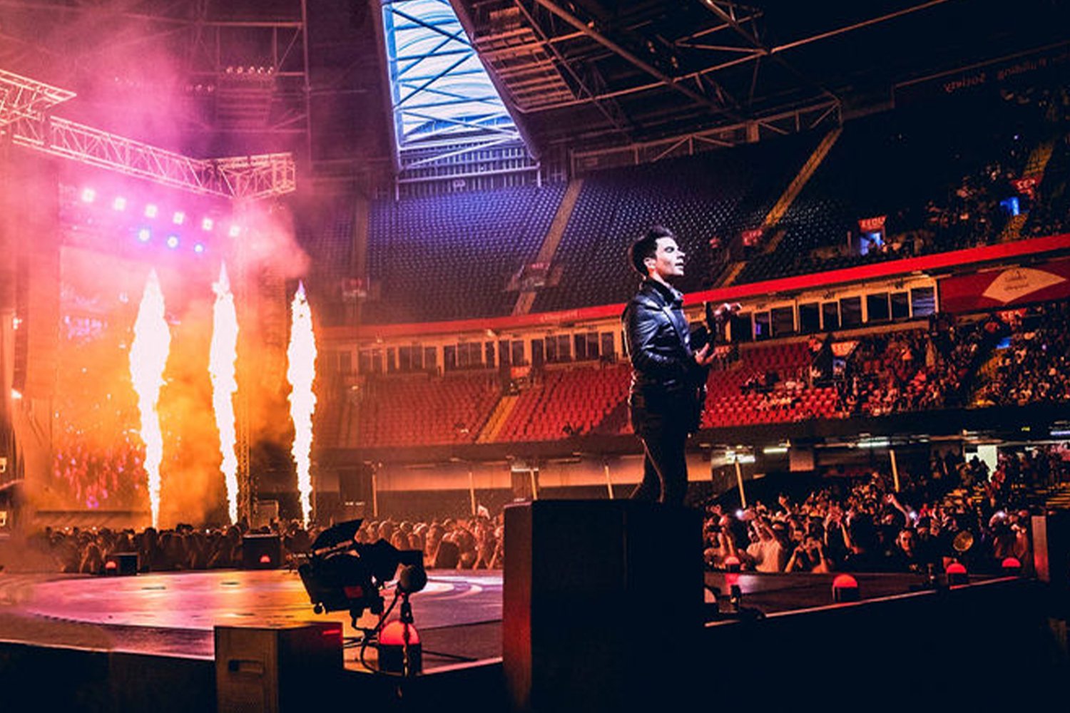 A performer standing on stage in an arena with colorful pyrotechnics and bright lights, with a large audience seated in the background.