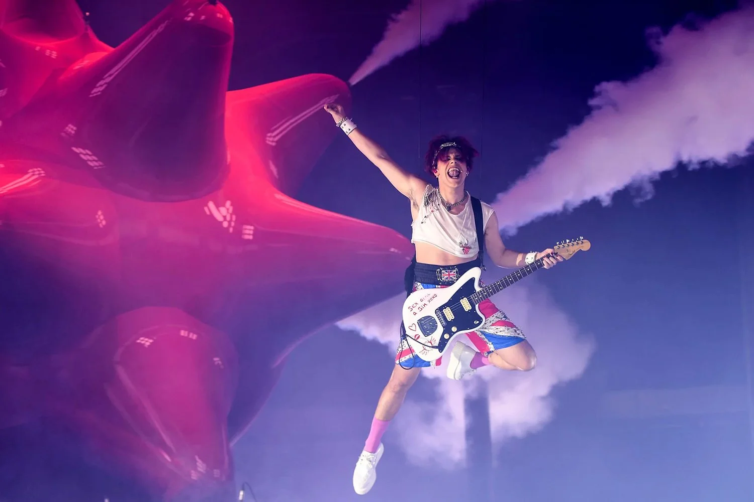 A woman jumping in the air playing an electric guitar with a large helium balloon and smoke in the background during a concert at night.