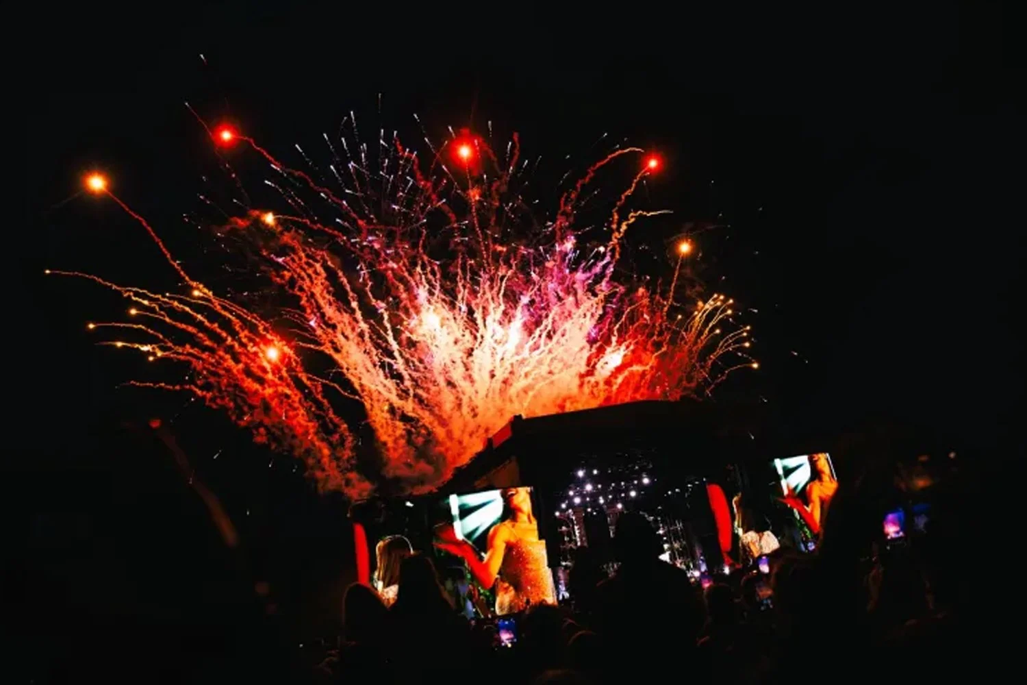 Fireworks lighting up the night sky above a concert stage with screens showing performers and an audience watching.