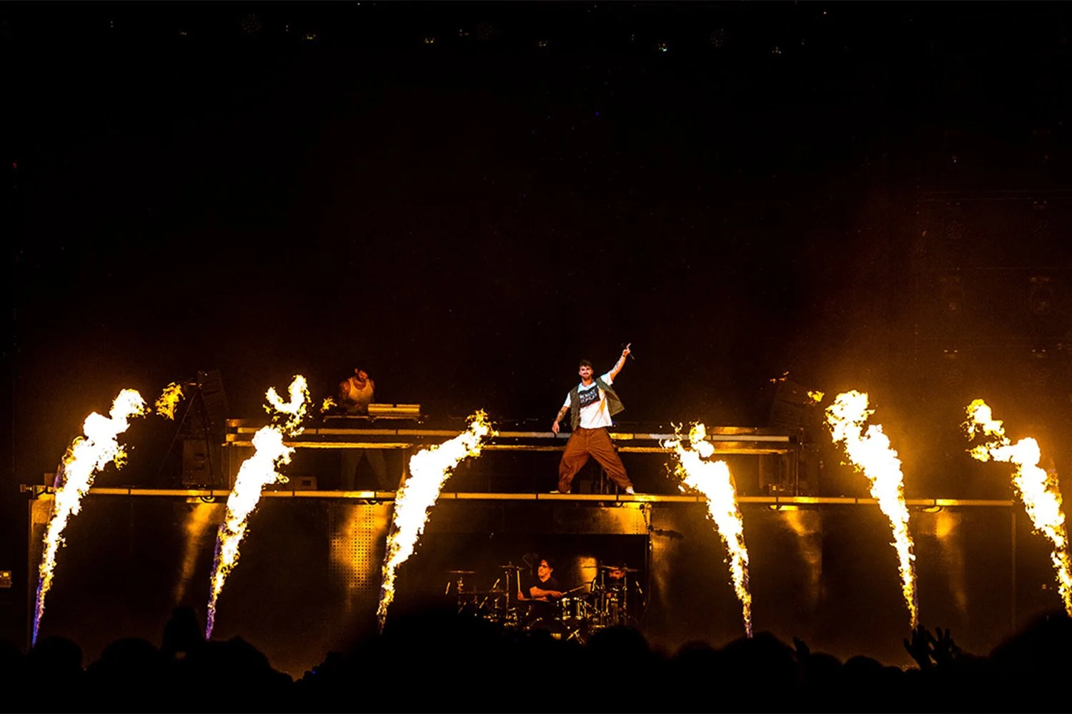 A live concert performance with a male singer standing on an elevated stage surrounded by fire bursts. The singer is mid-dance with one arm raised, wearing a patterned jacket and brown pants. Behind him, a DJ and a drummer are visible, with a dark background and audience silhouettes at the bottom of the image.