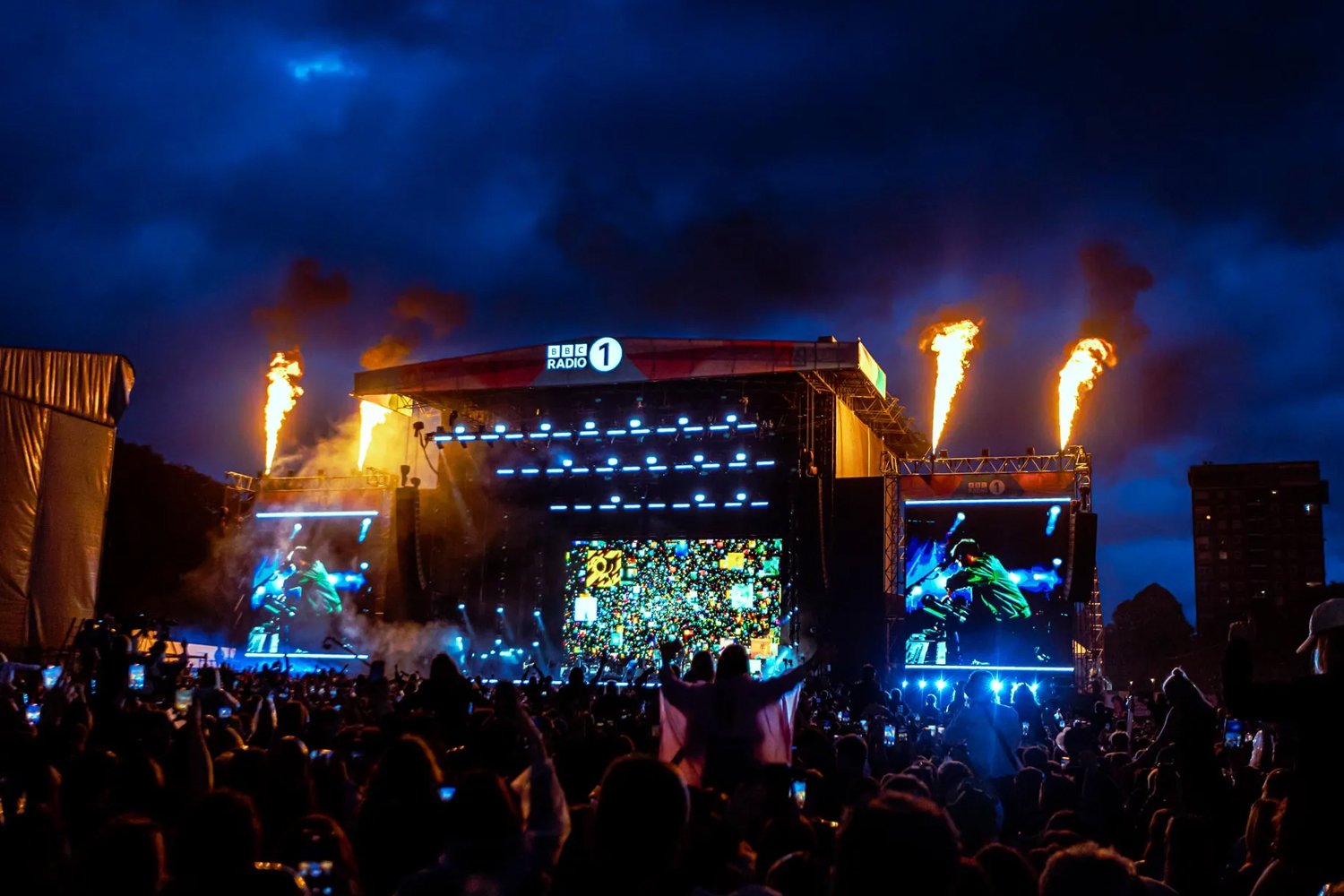 Crowd watching a concert at an outdoor stage with bright lights, digital screens, and fire effects against a dark evening sky.