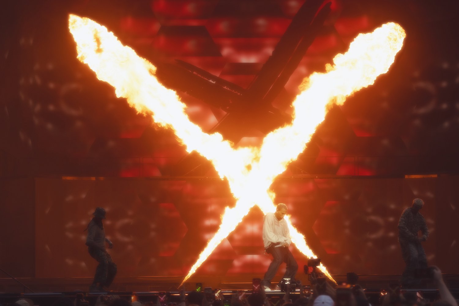 A concert stage with flames and fire effects, featuring three performers. One performer in the center stands near fire-looking projections, while the other two are positioned on either side. The background is illuminated in red and black tones.
