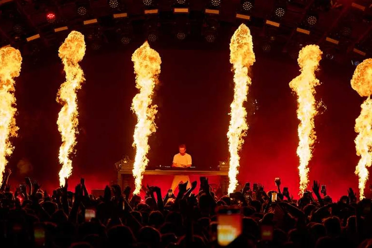 A night club scene with a DJ performing on stage, surrounded by flames shooting upward into the air, and a crowd of people watching and taking photos.