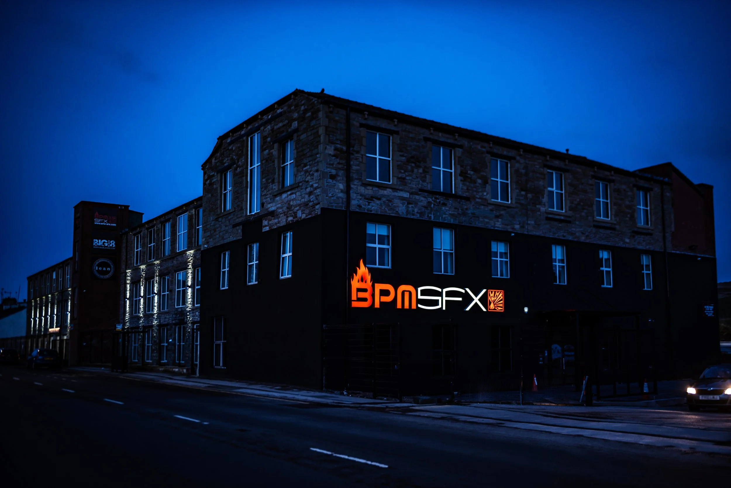Nighttime city street scene with a multi-story building made of stone and brick. The building has large windows and an illuminated sign that reads 'BPM SFX.' The sky is dark blue, and the street is mostly empty with a few cars visible.