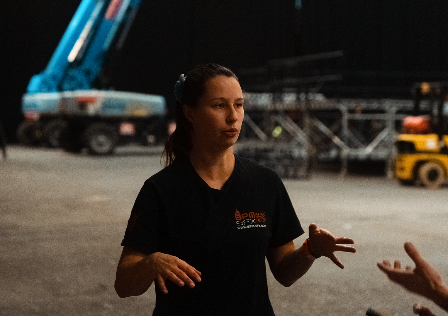 A woman with brown hair in a ponytail, wearing a black shirt with a logo that says BPM SFX, appears to be explaining or talking to someone in an industrial or construction setting at night. There is a blue crane and other heavy equipment in the background.