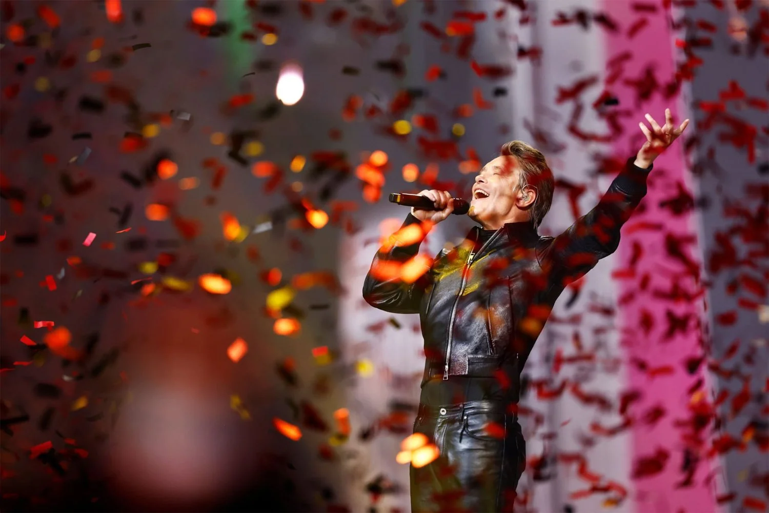 A person singing on stage surrounded by falling red, orange, and gold confetti, with a pink and white background.