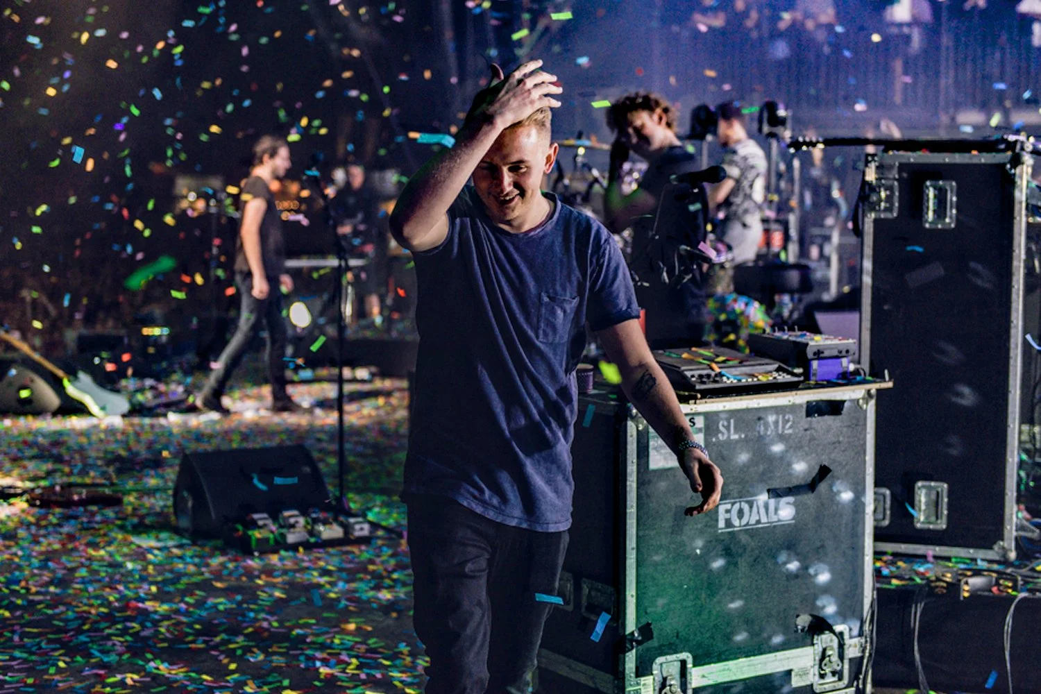 DJ smiling and adjusting his hair on a stage with colorful confetti falling, music equipment, and other people in the background.