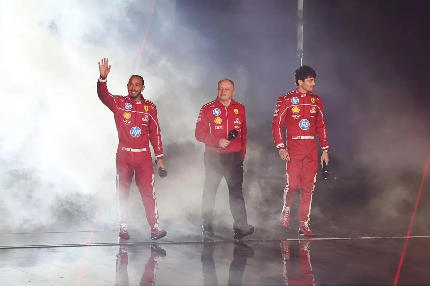 Three Ferrari Formula 1 drivers standing on a stage with smoke and lighting effects. The two drivers on the left and right are wearing red racing suits with sponsor logos, while the person in the middle is wearing a red jacket and holding a microphone. The driver on the left is waving to the crowd.