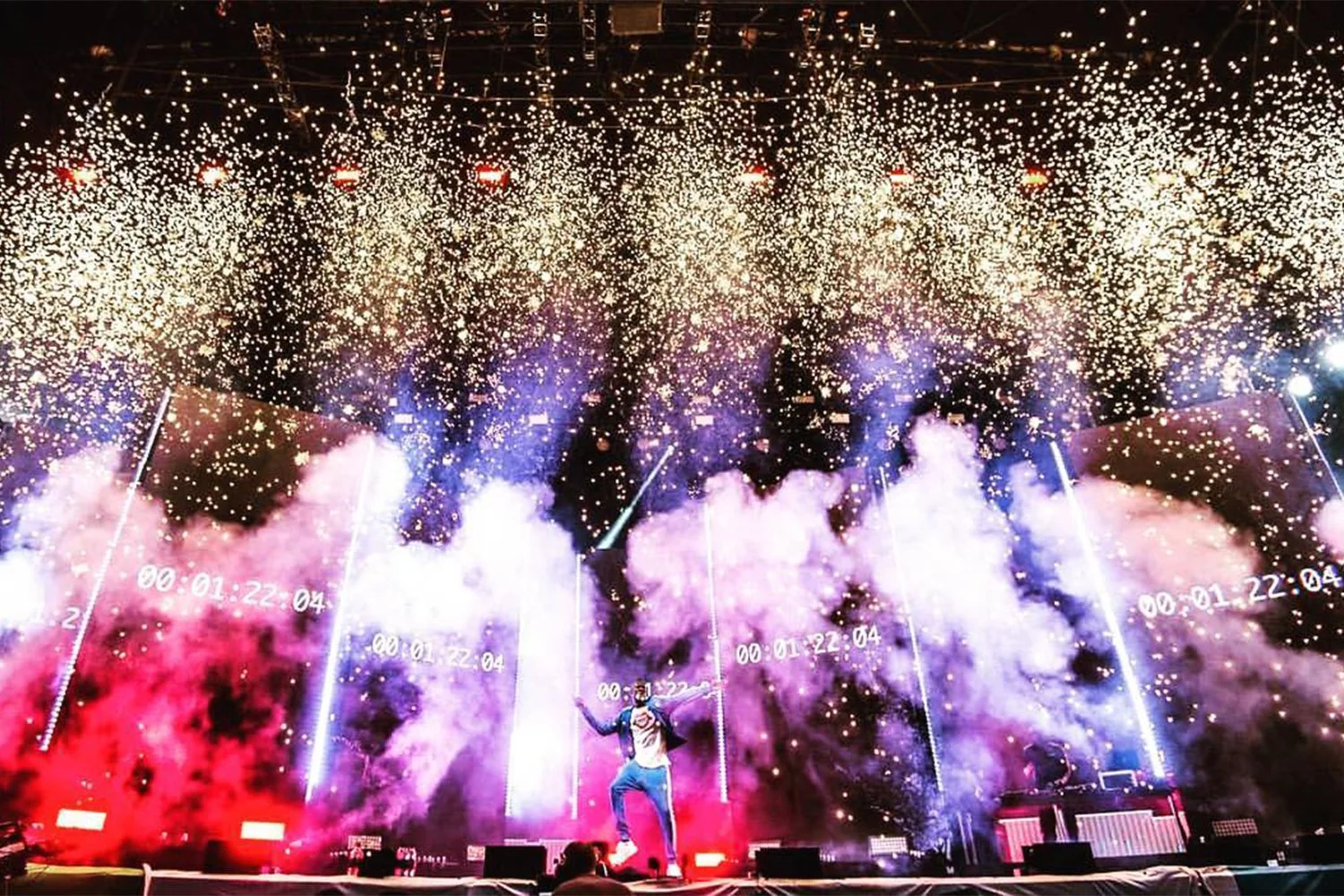 A performer stands on a stage with bright colors, smoke effects, and fireworks in the background during a concert or celebration.