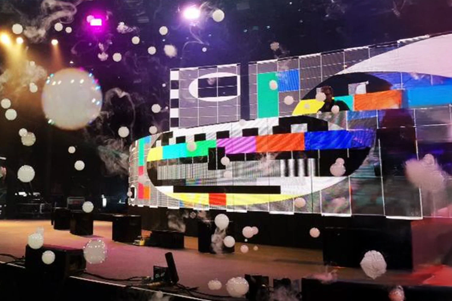 An electronic music stage with colorful lights and digital screens displaying abstract graphics, surrounded by white spherical balloons and fog.