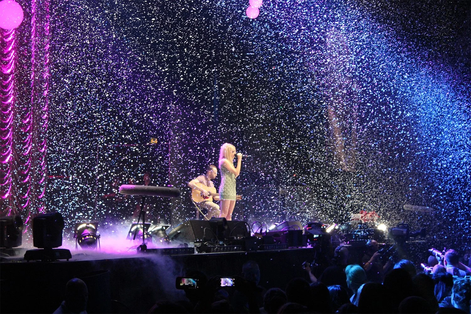 A singer and a guitarist performing on stage with colorful lighting and confetti falling from above, surrounded by an audience