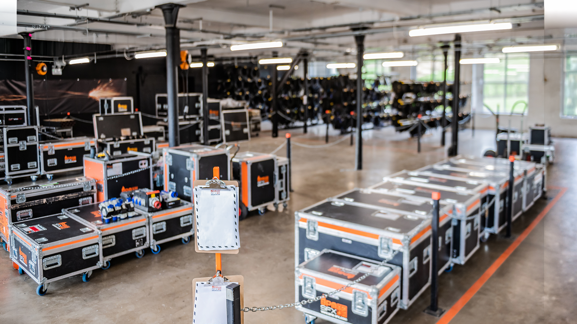 Empty equipment storage room with stacked black cases and equipment carts, possibly for event production or technical setup, with some cases on wheels and others on shelves, in a well-lit spacious area.