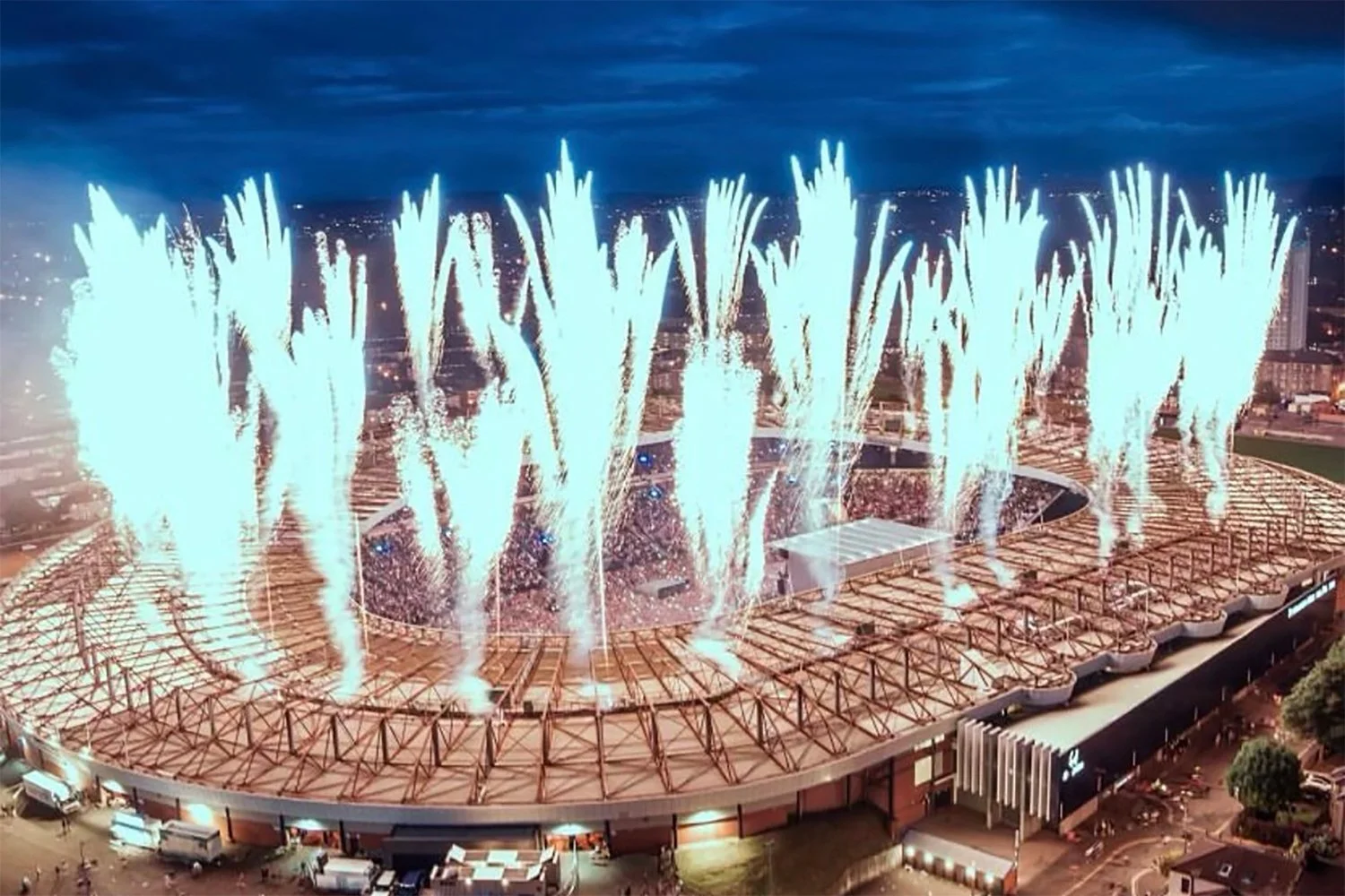 Fireworks display over a stadium at night, with bright white fireworks lighting up the sky and the stadium structure visible below.