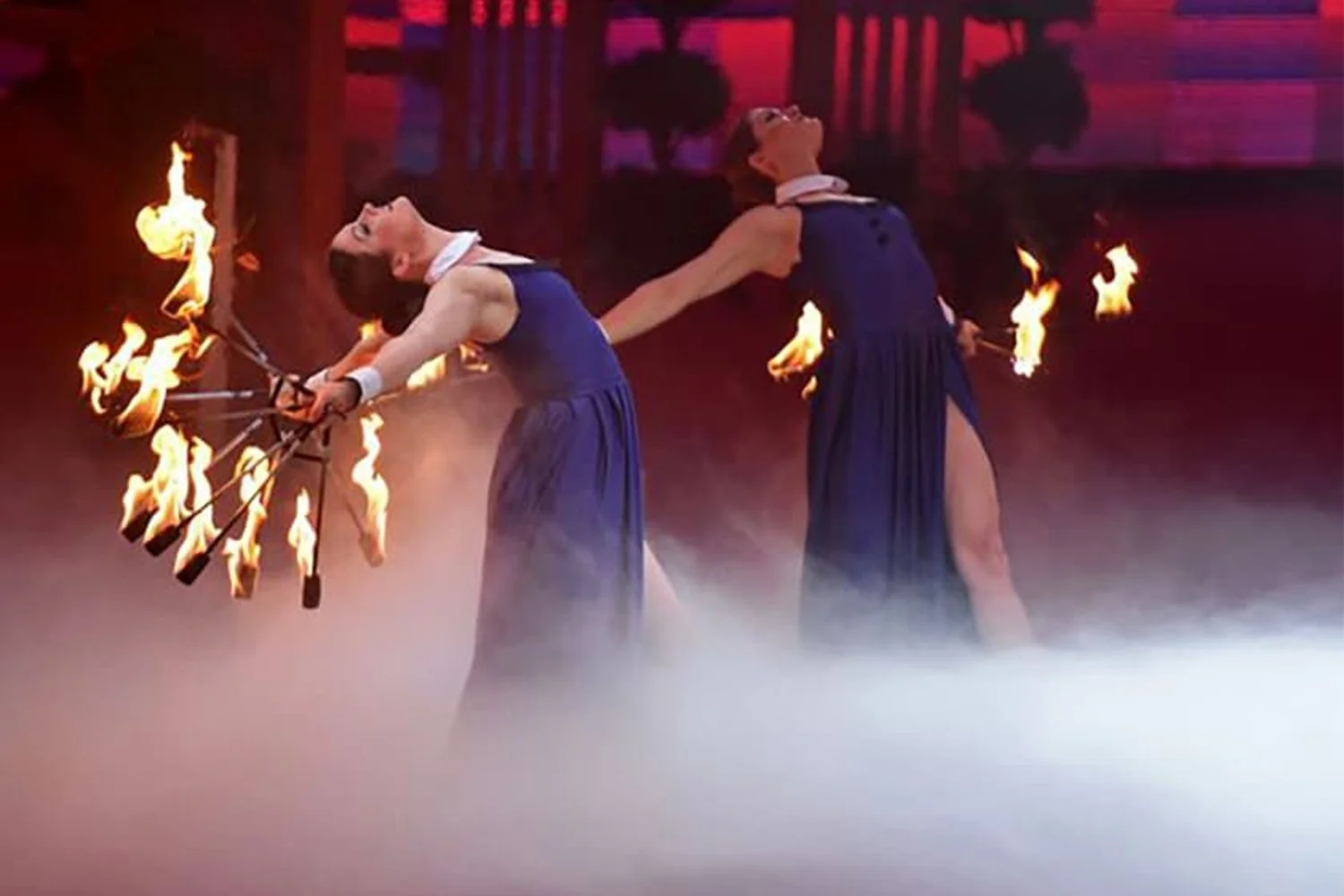Two female performers in blue dresses perform a fire-eating act on stage, with flames coming from metal rods they hold, amid fog and red lighting background.